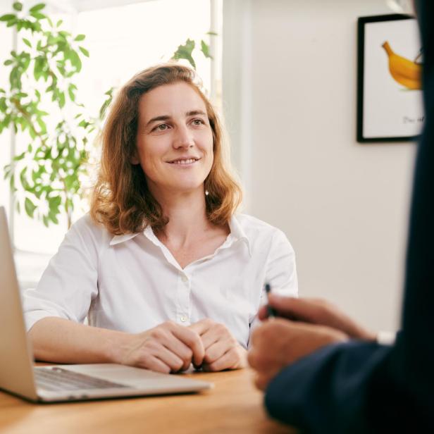Frau mit weißem Hemd sitzt an einem Holztisch vor einem Laptop, im Hintergrund eine grüne Pflanze und ein Bild mit einer Banane an der Wand.