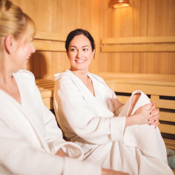 Zwei Frauen im Bademantel sitzen entspannt in einer Sauna mit Holzverkleidung und unterhalten sich.
