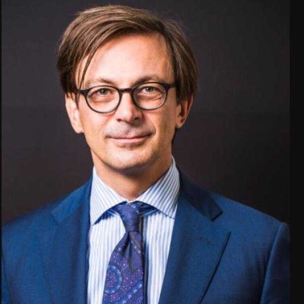 Ein Mann mit Brille, Anzug und Krawatte vor einem dunklen Hintergrund.