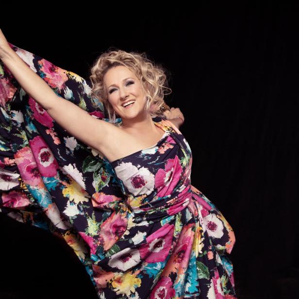 Eine Frau weht mit ihrem Kleid mit Blumenaufdruck und lächelt in die Kamera.