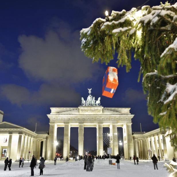 Brandenburger Tor bei Nacht mit Schnee, davor Menschen und ein Weihnachtsbaum mit goldener Kugel im Vordergrund.
