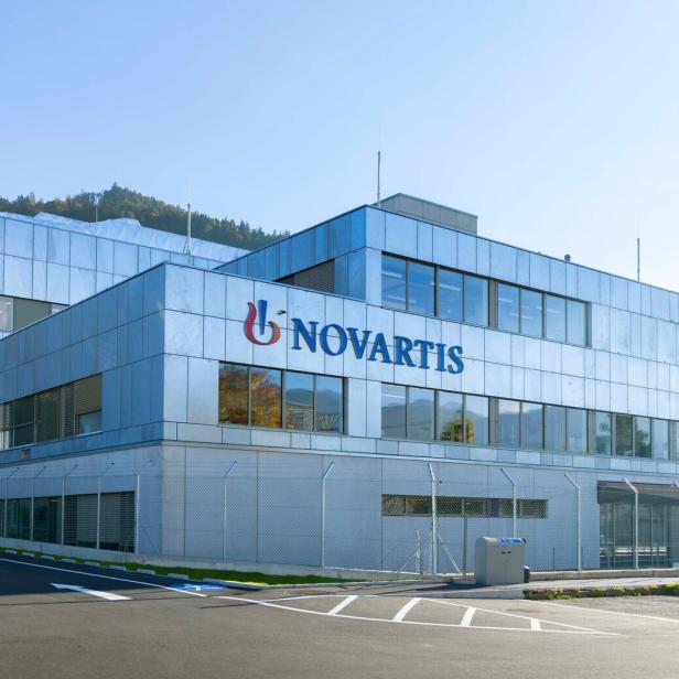 Modernes Bürogebäude mit Glasfassade und dem Schriftzug 'Novartis' an der Frontseite, umgeben von Parkplätzen und Bergen im Hintergrund.