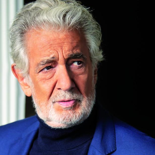 Älterer Mann mit grauen Haaren und Bart trägt einen dunkelblauen Anzug und schwarzen Rollkragenpullover vor schwarzem Hintergrund.