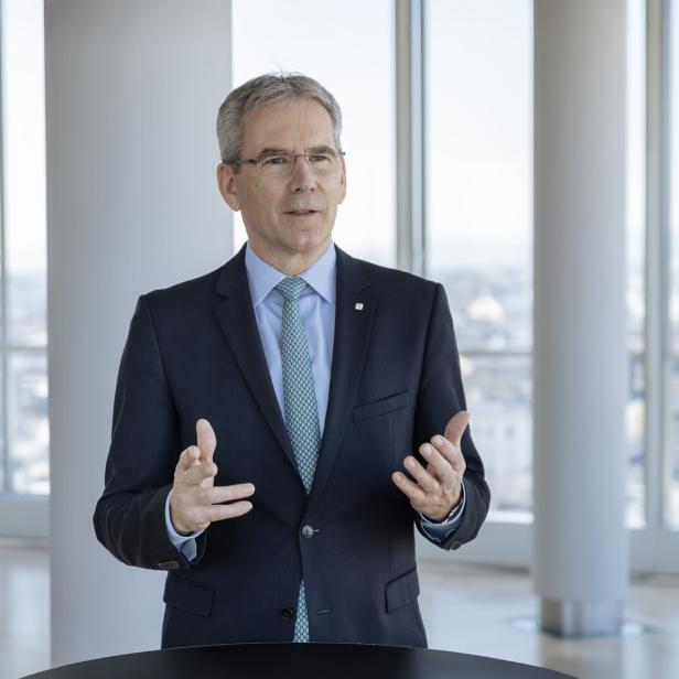 Mann in dunklem Anzug mit hellblauem Hemd und gemusterter Krawatte steht vor großen Fenstern mit Stadtblick, gestikuliert mit beiden Händen.