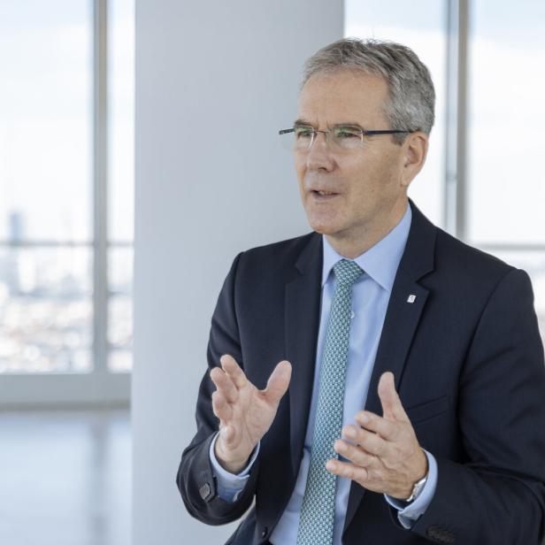 Mann in dunklem Anzug, hellblauem Hemd und gemusterter Krawatte sitzt vor großen Fenstern mit Stadtblick und gestikuliert mit den Händen.