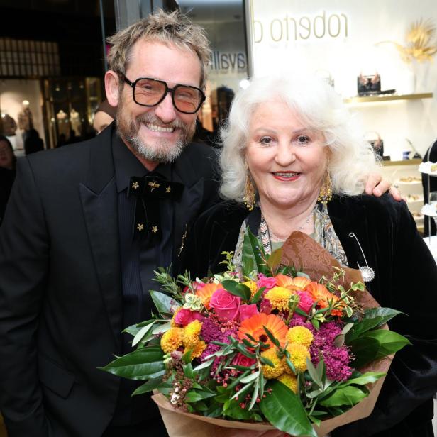 Zwei Personen lächeln, eine hält einen bunten Blumenstrauß, im Hintergrund ein Buffet.