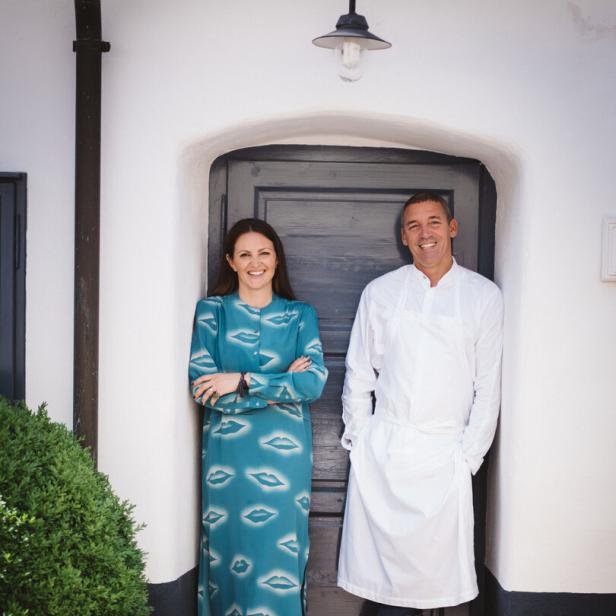 Eine Frau im türkisfarbenen Kleid und ein Mann in Kochkleidung stehen lächelnd nebeneinander vor einer dunklen Tür.