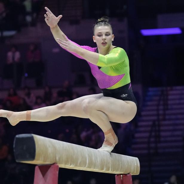 Eine Turnerin balanciert konzentriert auf einem Schwebebalken und führt eine Übung mit ausgestreckten Armen aus.