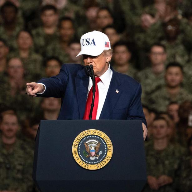 Donald Trump spricht vor einer Menschenmenge in Uniform, eine USA-Kappe tragend und auf etwas deutend.