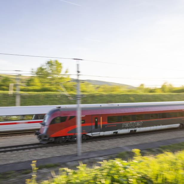 Zwei moderne Züge fahren nebeneinander auf parallelen Gleisen durch eine grüne Landschaft bei Sonnenschein.