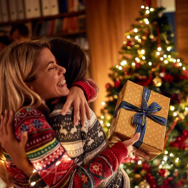 Zwei Frauen umarmen sich und übergeben sich Geschenke vor dem Christbaum in einem weihnachtlich geschmückten Raum.