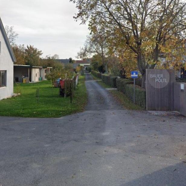 Eine schmale Straße führt zwischen einem weißen Haus und einem Holzzaun mit dem Schild „Bauer Pöltl“ in eine ländliche Gegend.