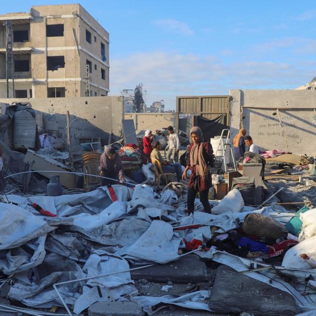 Mehrere Menschen durchsuchen Trümmer und Habseligkeiten vor zerstörten Gebäuden in einer städtischen Ruinenlandschaft.
