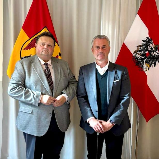 Zwei Männer im Anzug stehen nebeneinander vor einer rot-gelben und einer rot-weiß-roten Fahne mit Adler.