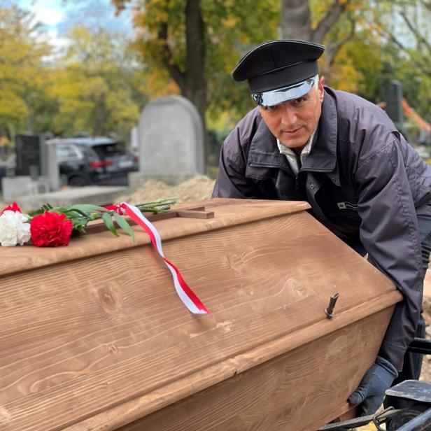 Ein Totengräber hebt einen Holzsarg mit Blumenschmuck in ein Grab.