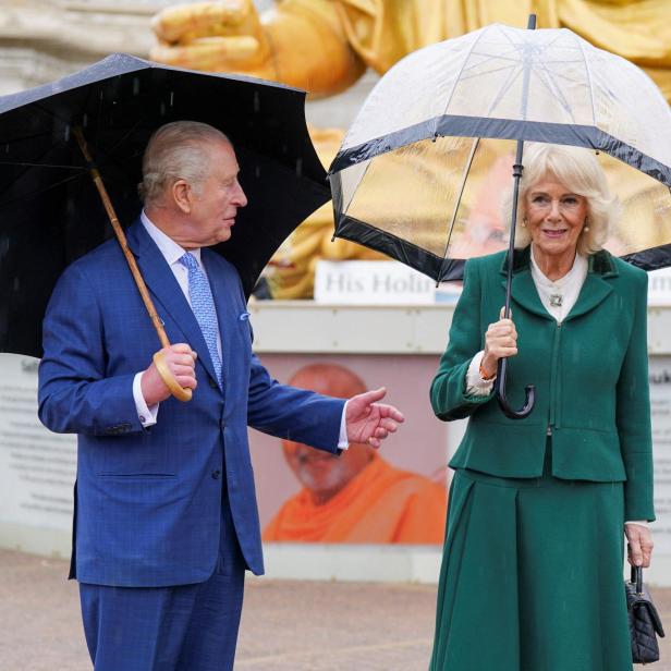 König Charles und Königin Camilla stehen im Regen unter Schirmen vor einem Denkmal.