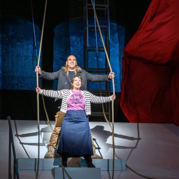 Zwei Personen stehen auf einer Bühne, halten Seile und lächeln, während im Hintergrund ein großes rotes Tuch hängt.