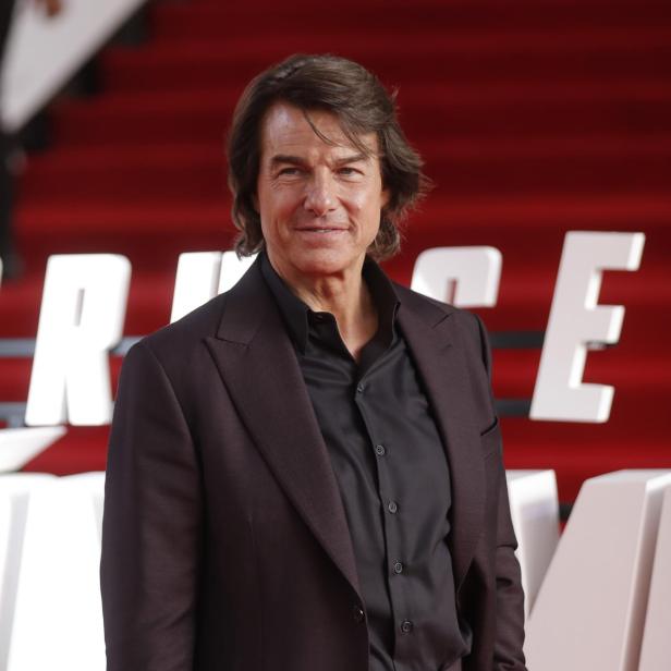 Tom Cruise posiert in einem dunklen Anzug vor einem roten Hintergrund mit weißen Buchstaben.