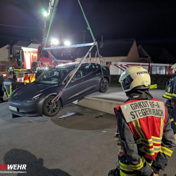 Ein Auto wird nachts von der Feuerwehr mit einem Kran angehoben, mehrere Feuerwehrleute beobachten den Einsatz.