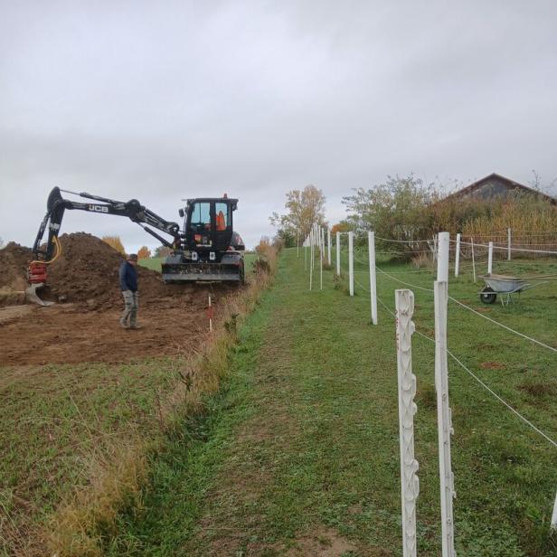 Ein Bagger hebt Erde aus, während ein Mann daneben steht und ein eingezäunter Grasweg vorbeiführt.