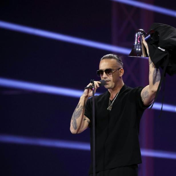 Raf Camora mit Sonnenbrille singt ins Mikrofon und hält einen Pokal hoch.