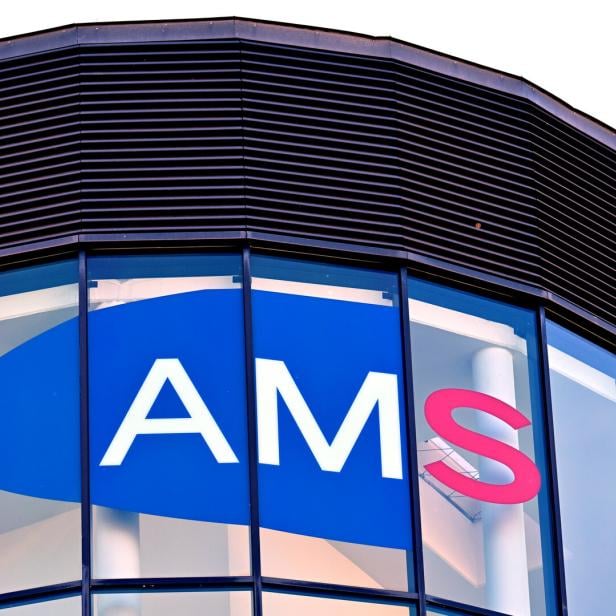 Großes AMS-Logo auf einer Glasfassade mit blauer und roter Schrift, darüber schwarze Lamellen.