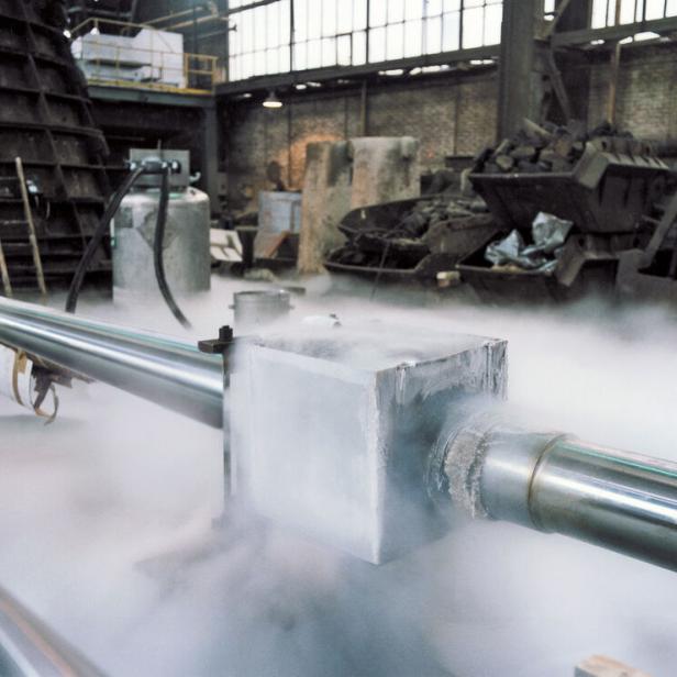 Metallrohre mit frostbedecktem Verbindungsstück, aus dem kalter Dampf entweicht, in einer Industriehalle.