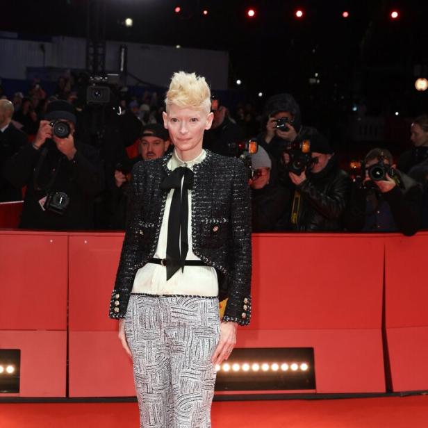 Eine Person mit kurzem, blondem Haar trägt eine gemusterte Hose, eine helle Bluse mit schwarzer Schleife und eine schwarze Jacke auf rotem Teppich.