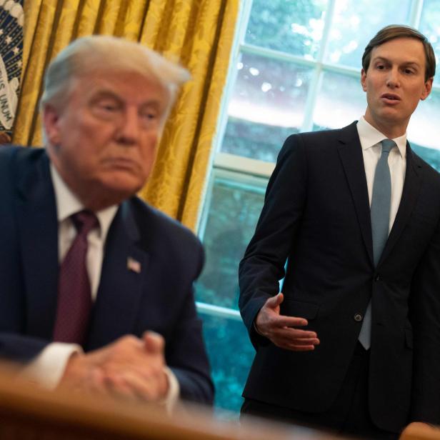 Donald Trump sitzt am Schreibtisch, während Jared Kushner im Anzug spricht.