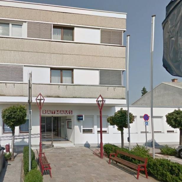 Ein modernes Rathaus mit flachem Dach, davor Bänke, Bäume, Fahnenmasten und geparkte Autos.