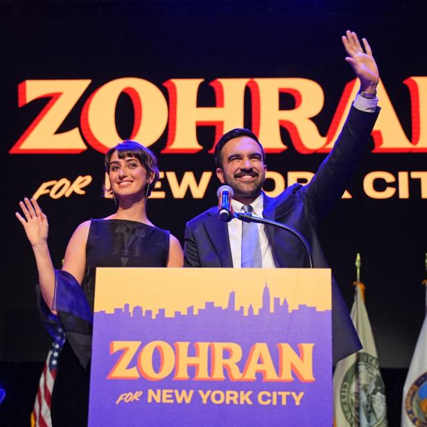 Zwei Personen, vermutlich Zohran und eine Frau, winken vor einem Wahlkampf-Banner für New York City.