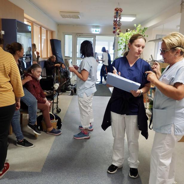 Mehrere Pflegekräfte und ältere Menschen befinden sich in einem Flur, einige sitzen, andere stehen und unterhalten sich.