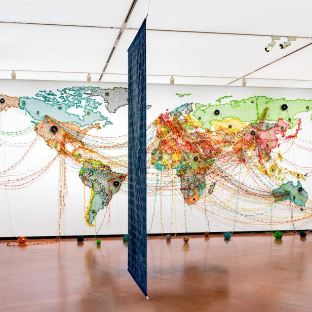 Weltkarte an der Wand mit farbigen Fäden, die verschiedene Punkte verbinden, und einem vertikal hängenden blauen Paneel in der Mitte des Raums.