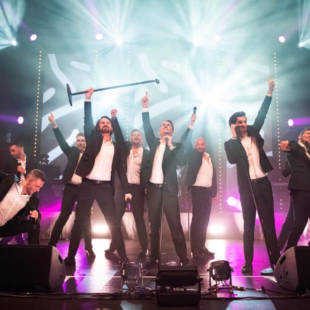 Zwölf Männer in schwarzen Anzügen und weißen Hemden stehen auf einer Bühne, einige mit Mikrofonen, andere mit erhobenen Armen, umgeben von Bühnenlicht und Nebel.