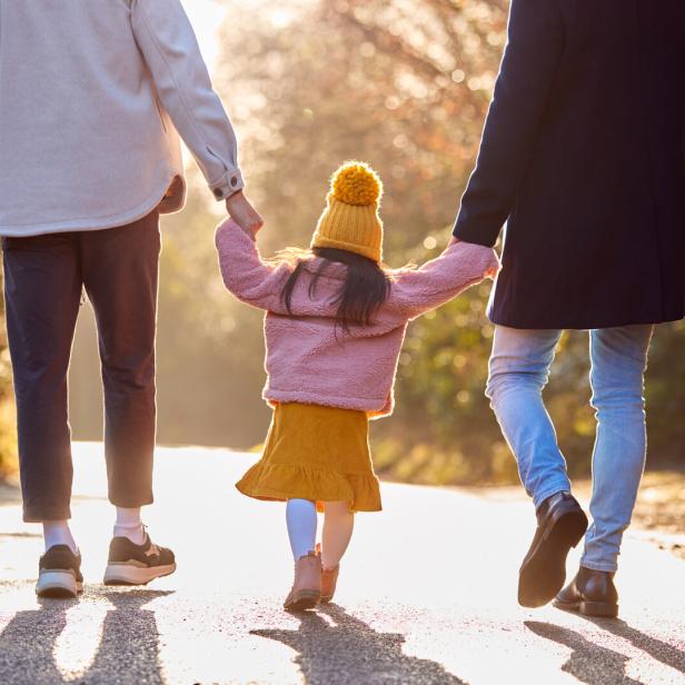 Ein Kind in gelber Mütze hält die Hände zweier Erwachsener und spaziert mit ihnen auf einem sonnigen Weg.