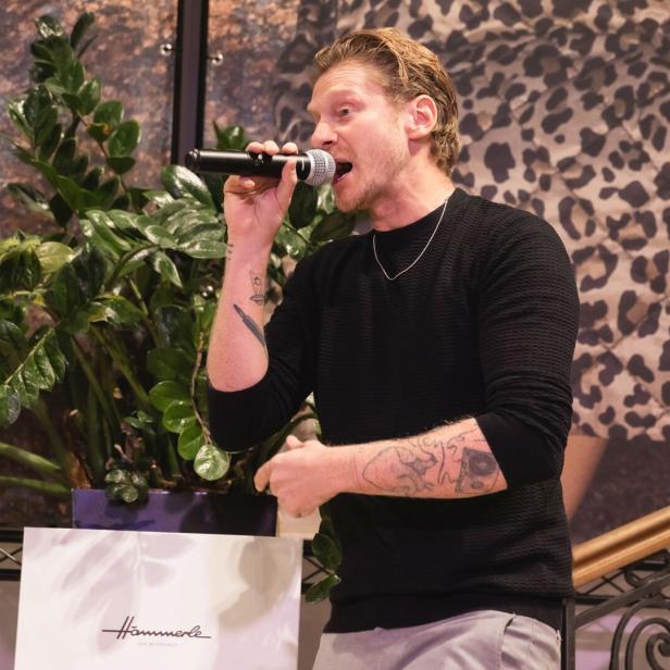 Ein Mann mit Tattoos singt mit einem Mikrofon, im Hintergrund sind grüne Pflanzen und ein Muster mit Leopardenflecken zu sehen.