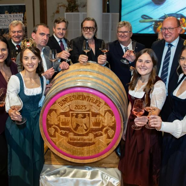 Mehrere Menschen stoßen feierlich mit Weingläsern um ein großes Weinfass mit der Aufschrift „Bundesweintaufe 2025“ an.