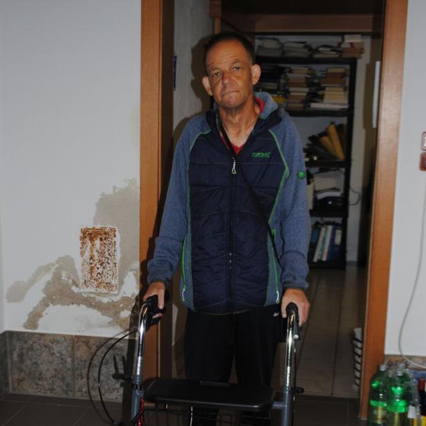 Ein Mann steht mit einem Rollator in einem Flur, im Hintergrund sind Regale mit Akten und Getränkeflaschen zu sehen.