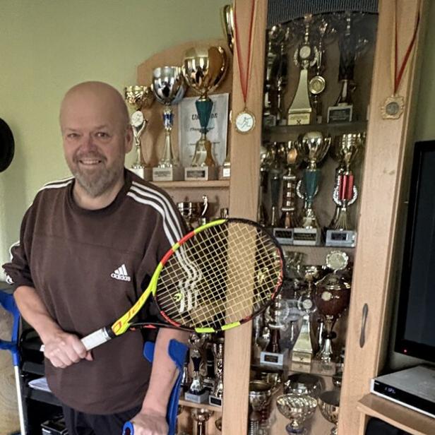 Ein Mann mit Beinprothese und Krücken hält einen Tennisschläger und steht vor einer Vitrine voller Pokale.