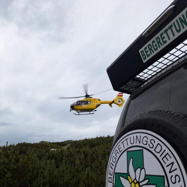 Ein gelber Rettungshubschrauber fliegt über eine Berglandschaft, im Vordergrund ein Fahrzeug der Bergrettung Österreich.