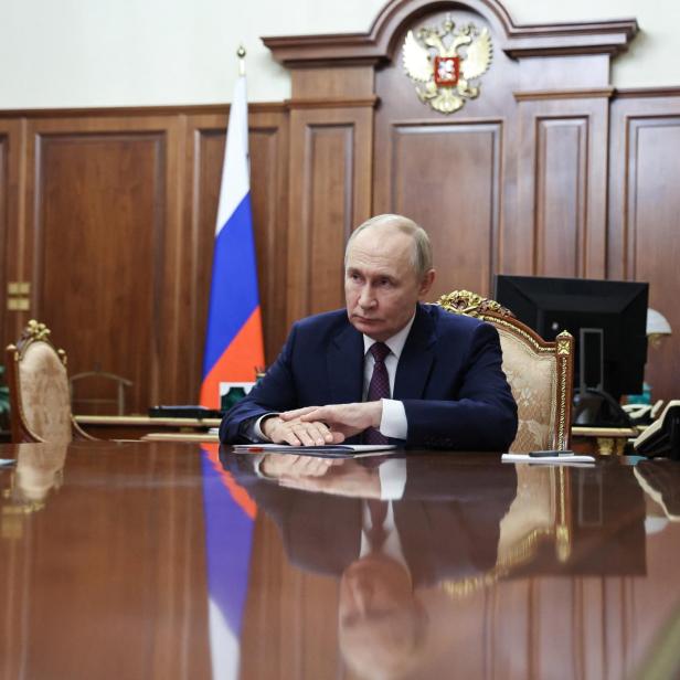 Putin sitzt an einem polierten Holztisch vor russischen Flaggen und dem Staatswappen.