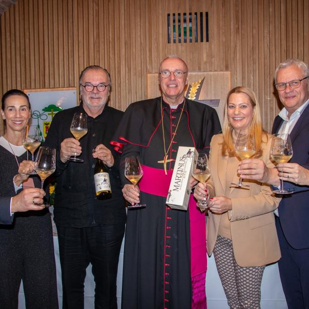 Sieben Personen stoßen mit Weißweingläsern an, eine Person trägt eine Bischofskleidung und hält eine Geschenkverpackung.