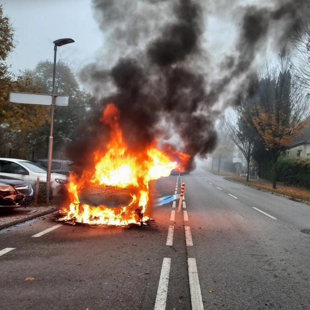 Ein Auto steht auf einer Straße in Flammen, dichter schwarzer Rauch steigt auf.