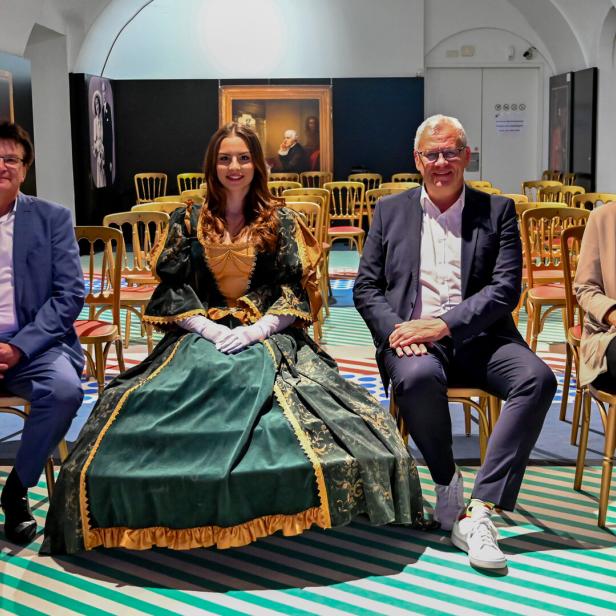 Vier Personen sitzen nebeneinander auf Stühlen, eine Frau trägt ein historisches Kleid, im Hintergrund sind Gemälde an den Wänden.