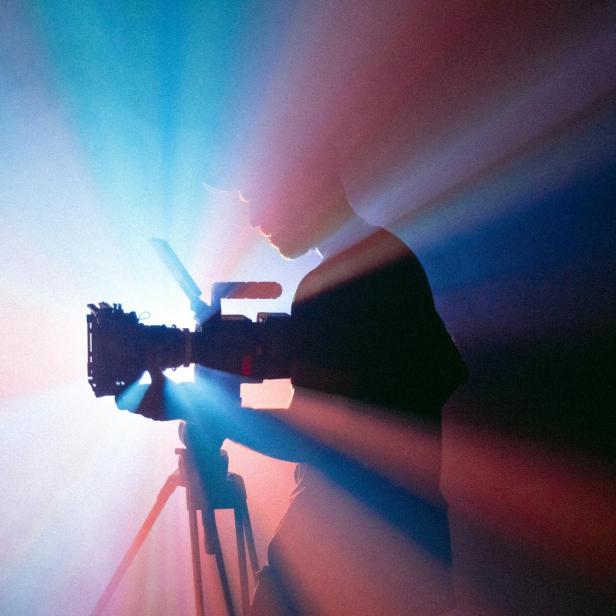 Silhouette einer Person, die eine Kamera auf einem Stativ bedient, umgeben von blau-rotem Lichtschein.