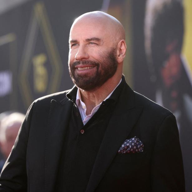 John Travolta lächelt mit Bart und Anzug vor einem unscharfen Hintergrund.