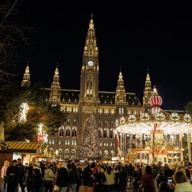 Festlich beleuchteter Weihnachtsmarkt mit Karussell und vielen Menschen vor einem großen historischen Gebäude bei Nacht.
