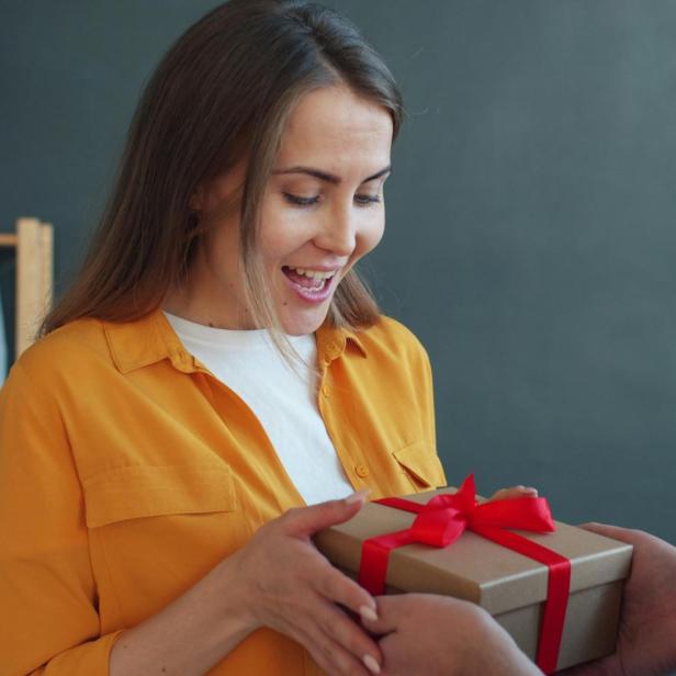 Frau in gelbem Hemd nimmt Geschenk mit roter Schleife entgegen, Mann reicht es ihr.
