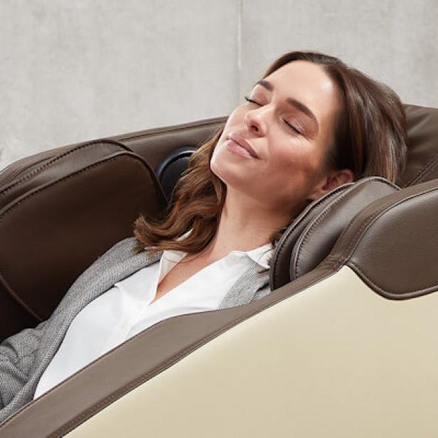 Frau mit braunen Haaren lehnt entspannt in einem braun-beigen Massagesessel, trägt grauen Blazer und weiße Bluse.