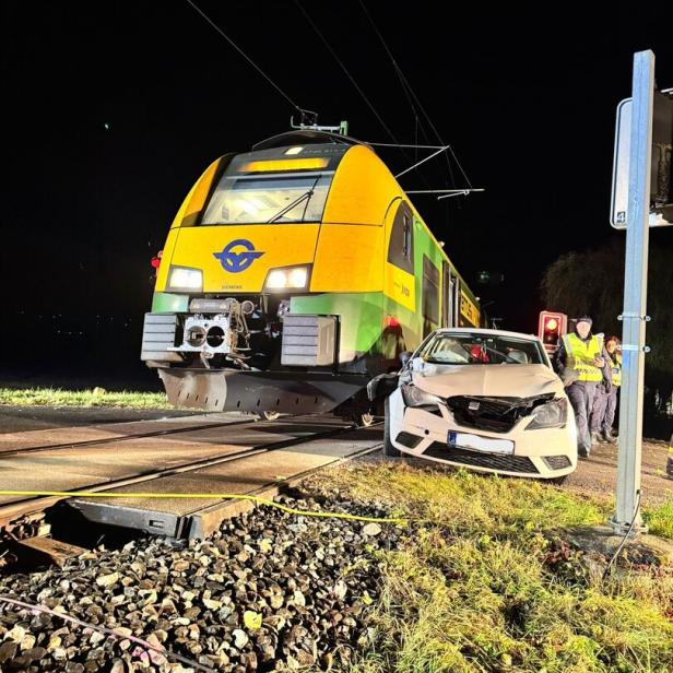 Ein Zug und ein beschädigtes Auto stehen nach einem Unfall auf einem Bahnübergang, mehrere Personen beobachten die Szene.